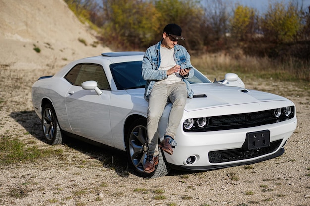 Knappe man in jeansjasje en pet zit op kap bij zijn witte muscle car in carrière en sms't op telefoon