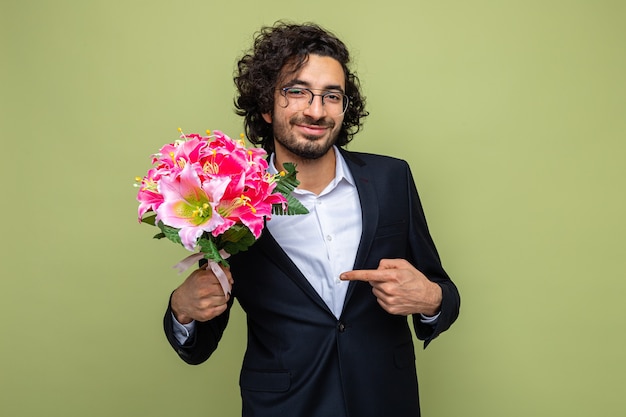 Knappe man in pak met boeket bloemen wijzend met wijsvinger naar glimlachend zelfverzekerd vieren