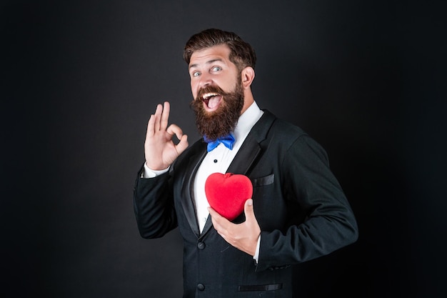 Knappe man in smoking met liefdesgeschenk van hart voor valentijnsdag laat ok gebaar valentijn zien