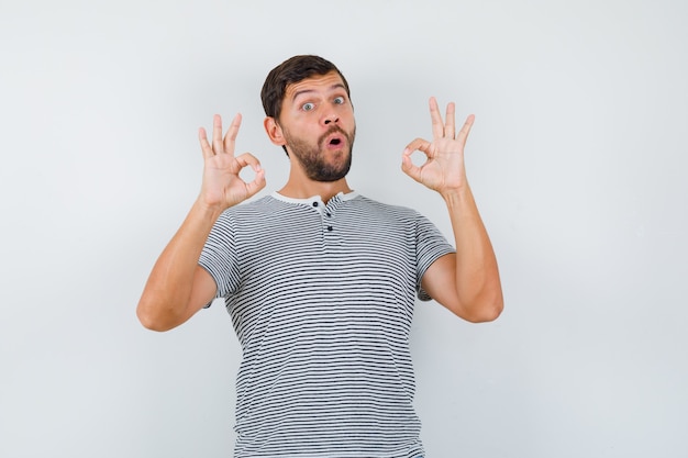 Knappe man in t-shirt met ok teken terwijl hij zijn mond opent en geschokt kijkt, vooraanzicht.