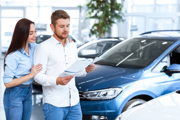 Knappe man leest een reclameboekje bij de autodealer terwijl zijn mooie vrouw omarmen