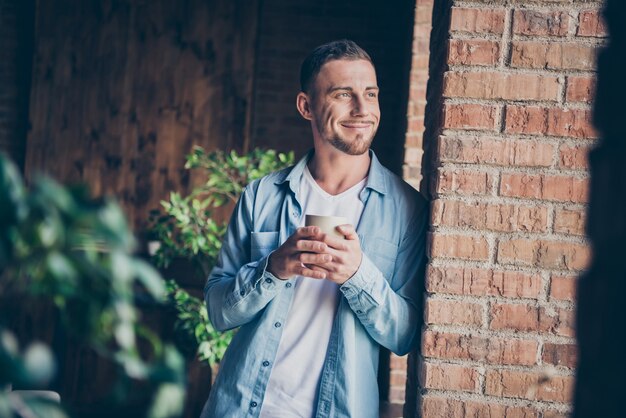 knappe man leunend bakstenen ontwerp muur kijken venster warme koffie drinken