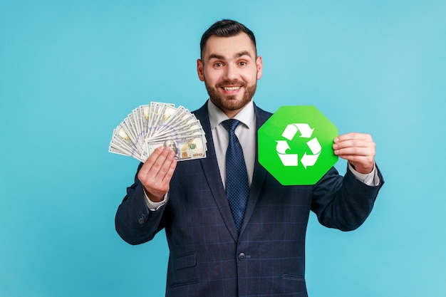 Knappe man met baard die een officieel pak draagt met een vrolijke uitdrukking die dollarbankbiljetten en groene recycling laat zien die naar de camera zingt. Indoor studio opname geïsoleerd op blauwe achtergrond.