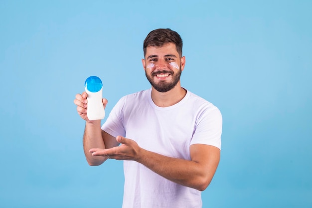 Knappe man met baard op vakantie in badkleding met fles zonnebrandcrème die er positief en gelukkig uitziet en glimlacht met een zelfverzekerde glimlach die zijn tanden laat zien