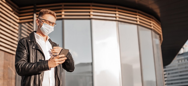 Knappe man met een beschermend masker op zijn gezicht met een bril met een smartphone op straat van een grote stad. Zakenman praten aan de telefoon op stedelijke achtergrond