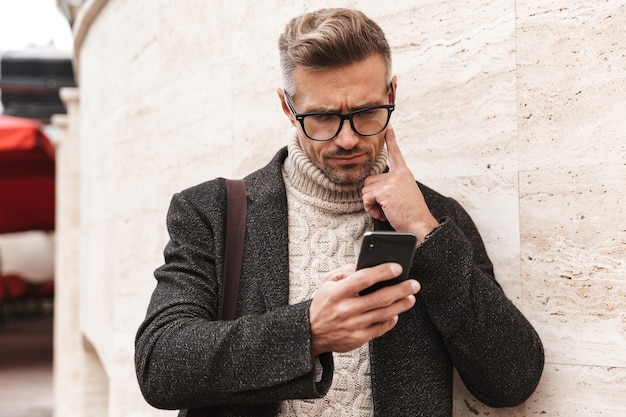 Knappe man met een jas buiten lopen, met mobiele telefoon