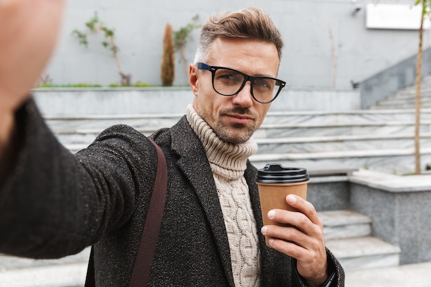 Knappe man met een jas buiten wandelen, een selfie nemen, met afhaalmaaltijden koffiekopje