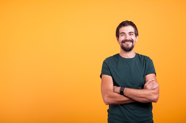 Knappe man met zijn armen gekruist glimlachen naar de camera geïsoleerd op gele achtergrond. Portret van aantrekkelijke bebaarde zelfverzekerde persoon in studiofoto