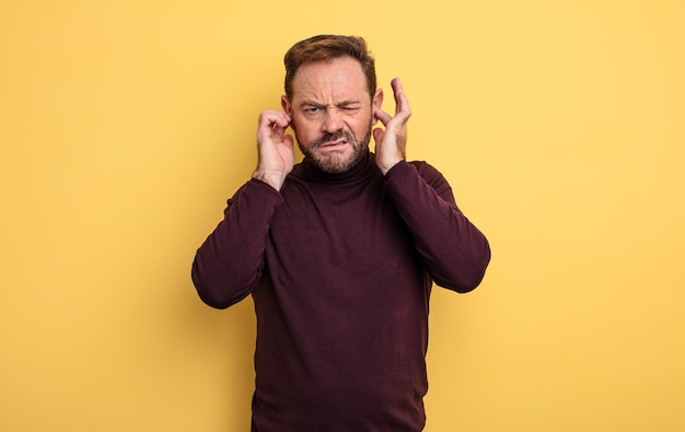 Foto knappe man van middelbare leeftijd die boos, gestrest en geïrriteerd kijkt en beide oren bedekt voor een oorverdovend geluid, geluid of luide muziek