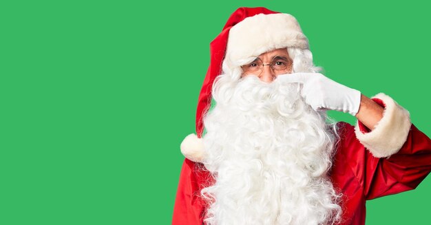 Knappe man van middelbare leeftijd met kerstmankostuum en baard staand Wijzend met handvinger naar gezicht en neus, vrolijk glimlachend. Schoonheidsconcept
