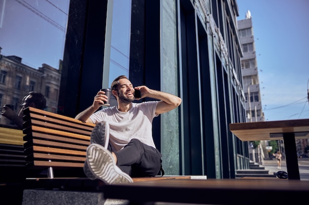 Knappe man zittend op een bankje met smartphone en koffie buiten