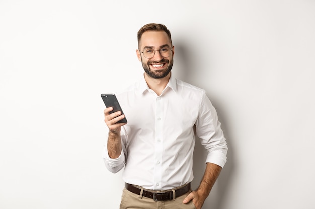 Knappe manager die smartphone gebruikt en tevreden glimlacht, sms-bericht verzendt, staande op een witte achtergrond