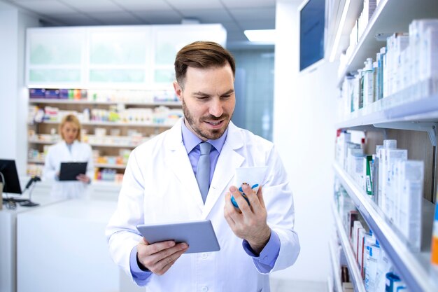 Knappe mannelijke apotheker die in witte jas geneeskunde in apotheekopslag of drogisterij houdt en prijs op tabletcomputer controleert.