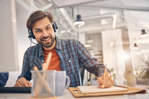 Knappe mannelijke werknemer in headset die naar de camera kijkt en glimlacht terwijl hij aan tafel zit en aantekeningen maakt op kantoor