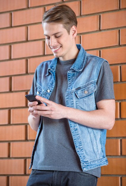 Knappe mens die bij bakstenen muur leunt en telefoon bekijkt.