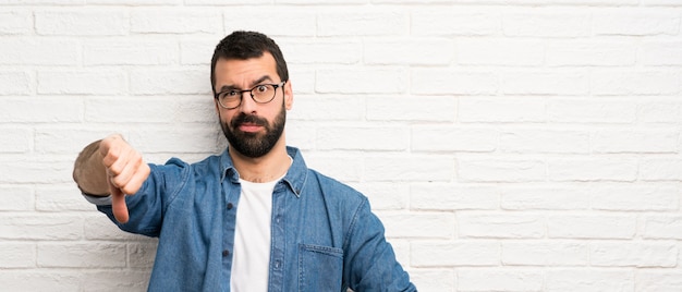 Knappe mens met baard over witte bakstenen muur die duim neer met negatieve uitdrukking tonen