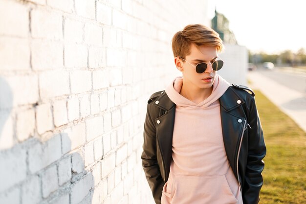 Knappe modieuze jonge hipster man in zwarte zonnebril in stijlvolle zwart lederen jas in een roze sweatshirt staande in de buurt van een witte bakstenen muur van de zonnestralen. aantrekkelijke kerel.