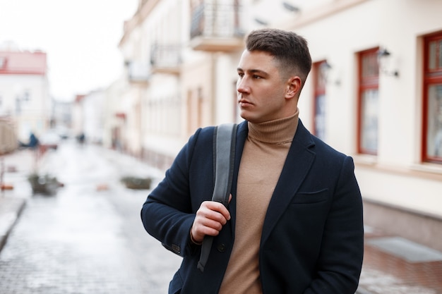 Knappe modieuze jongeman in een stijlvolle zwarte jas in een beige gebreide trui met een trendy rugzak met een stijlvol kapsel vormt in de stad