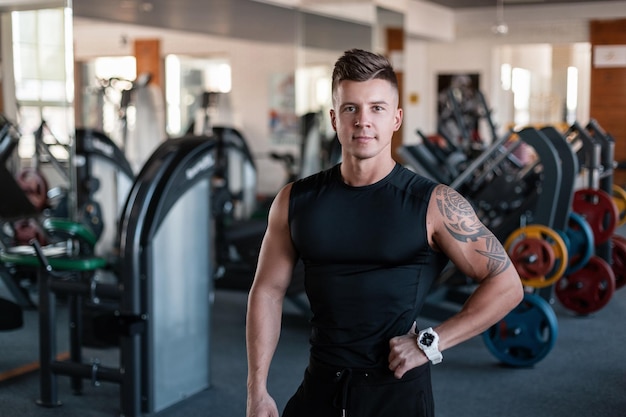 Knappe modieuze sportieve atleet bodybuilder man model met kapsel in zwarte sportkleding met zwarte tshirt staat en poseert in de sportschool