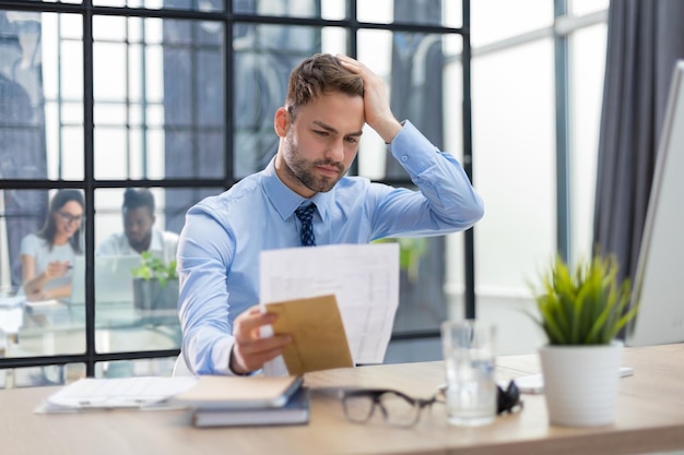 Knappe ondernemer leest een brief uit een envelop in een desktop op kantoor met collega's op de achtergrond