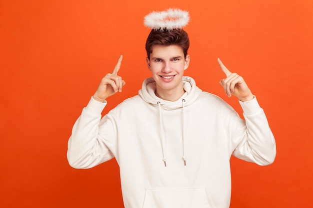 Knappe onschuldige jongeman in casual sweatshirt wijzende vingers op pluizige nimbus overhead en glimlachend, zegenend. Indoor studio-opname geïsoleerd op oranje achtergrond