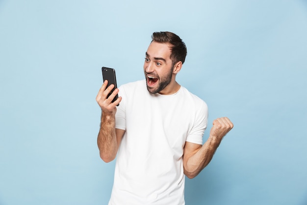 knappe opgewonden jonge man met behulp van mobiele telefoon geïsoleerd over blauwe muur maken winnaar gebaar.