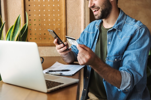 knappe optimistische man met een denim shirt met een mobiele telefoon en een creditcard met een laptop terwijl hij binnenshuis in een café werkt