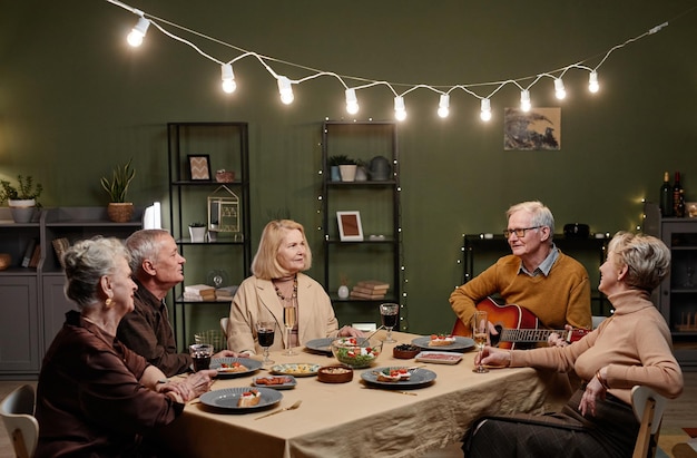 Knappe oude man die akoestische gitaar speelt terwijl hij aan de eettafel zit in een ingerichte woonkamer en een feestelijk diner heeft met zijn vier vrienden