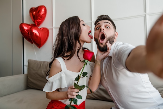 knappe paar man en vrouw maken selfie met rode roos en hartvormige ballonnen thuis op de bank