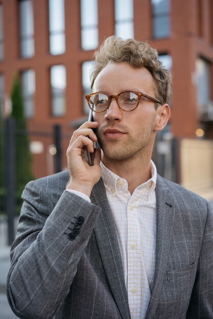 Knappe peinzende zakenman met een bril die op een mobiele telefoon praat die op straat staat