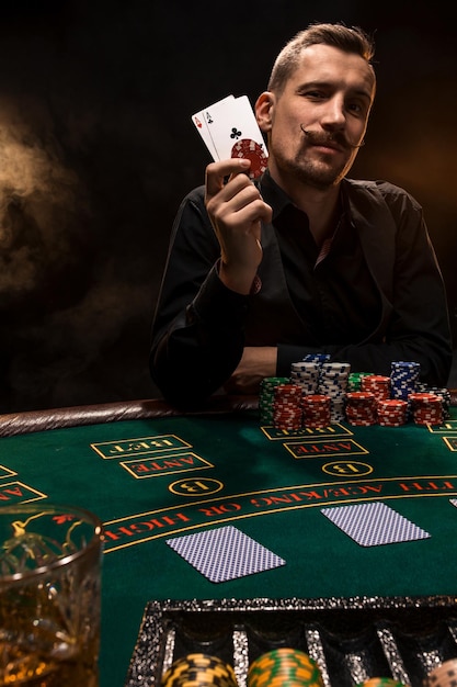 Knappe pokerspeler met twee azen in zijn handen en chips aan de pokertafel in een donkere kamer vol sigarettenrook.