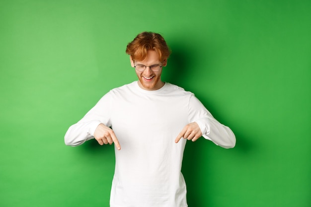 Foto knappe roodharige man met een bril en witte lange mouwen die naar een advertentie kijkt, met de vingers wijst en naar beneden kijkt met een tevreden glimlach, staande over een groene achtergrond