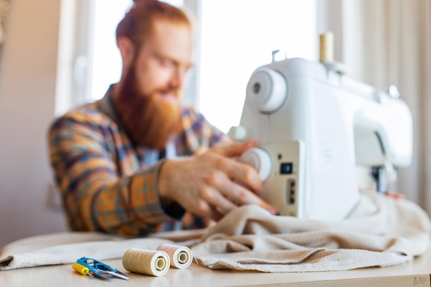 Knappe roodharige man met lange baard naait op een naaimachine in de thuisstudio