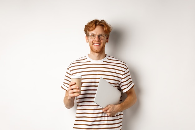 Knappe roodharige mannelijke werknemer in glazen die pauze heeft, koffie drinkt en laptop vasthoudt, staande op een witte achtergrond