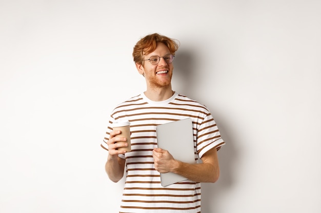 Knappe roodharige mannelijke werknemer in glazen met pauze, koffie drinken en laptop vasthouden, staande op een witte achtergrond.