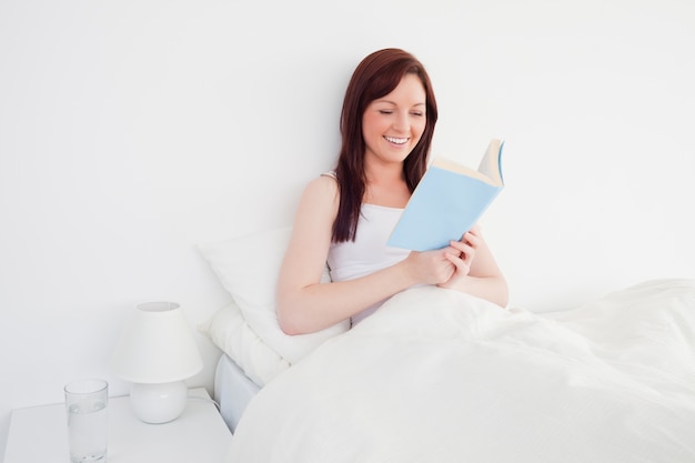 Knappe roodharige vrouw die een boek leest terwijl het zitten op haar bed