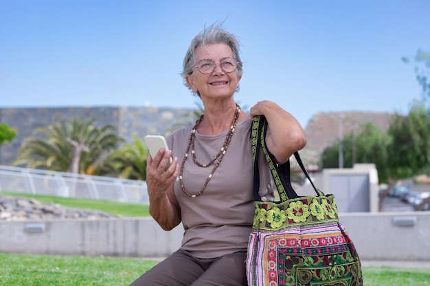 Knappe senior blanke vrouw die buiten in het park zit en een mobiele telefoon vasthoudt die een oudere dame vasthoudt die van vakantie geniet en ontspant