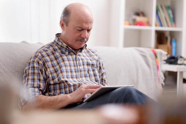 Knappe seniorman met behulp van een tablet thuis zittend op een bank