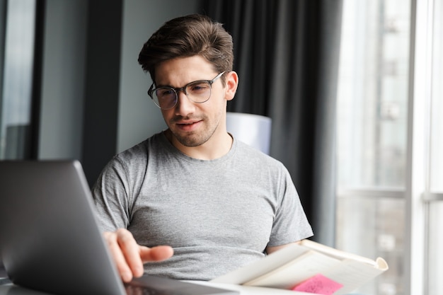 Knappe serieuze jonge bebaarde man die vrijetijdskleding draagt met een laptopcomputer terwijl hij thuis aan tafel zit