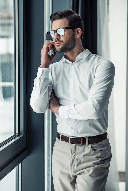 Knappe serieuze zakenman in bril praat op de mobiele telefoon terwijl hij uit het raam kijkt in kantoor