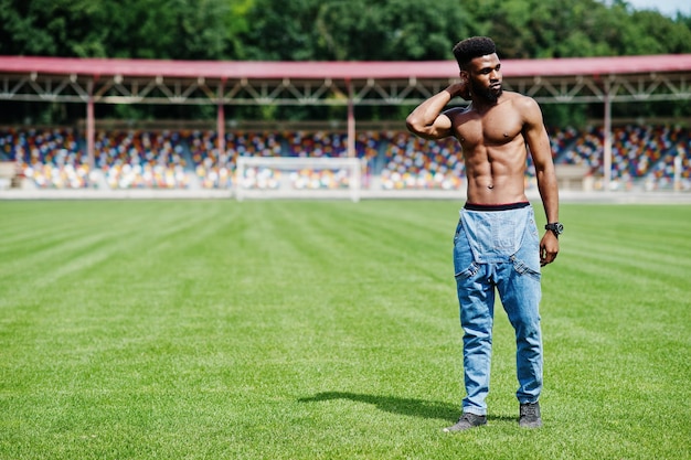 Knappe sexy Afro-Amerikaanse gespierde blote torso man op jeans overall gesteld op groen gras van stadion voetbalveld modieuze zwarte man portret