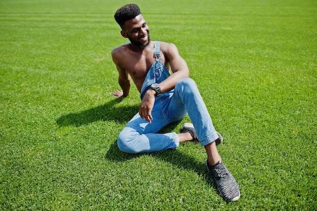 Knappe sexy Afro-Amerikaanse naakte torso man op jeans overall zittend op groen gras van stadion voetbalveld Modieuze zwarte man portret