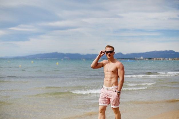 Knappe shirtless atletische jonge kerel die op het strand door de waterlijn loopt. jonge gespierde man met een perfect lichaam in korte broek en zonnebril wandelen door de kust. reismode. sport levensstijl