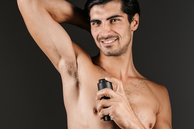 Knappe shirtless man permanent geïsoleerd, deodorant sproeien op zijn oksel