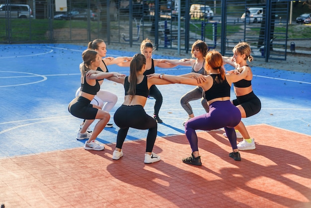 Knappe slanke atletiekvrouwen die hurkende oefeningen maken die in cirkel op het openluchtstadion staan in het moderne stadspark.