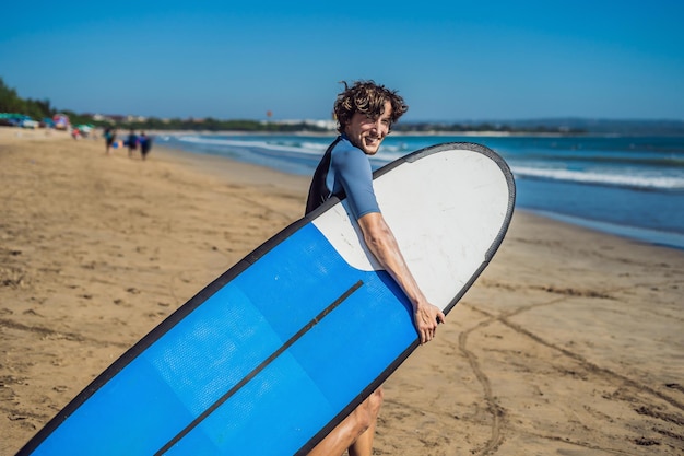 Knappe sportieve jonge surfer poseren met zijn surfplank onder zijn arm in zijn wetsuit op een tropisch zandstrand