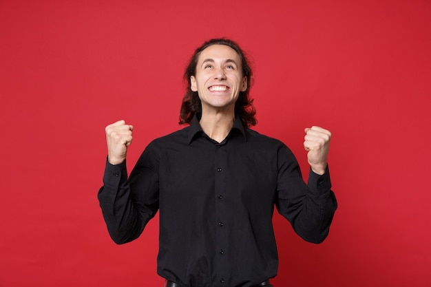 Knappe stijlvolle jonge krullend langharige man in zwart shirt poseren geïsoleerd op rode muur achtergrond studio portret. mensen oprechte emoties levensstijl concept. bespotten kopie ruimte. winnaar gebaar doen.