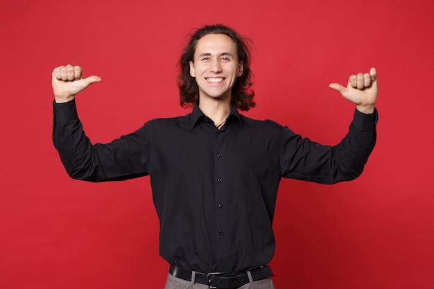 Knappe stijlvolle jonge krullend langharige man in zwart shirt poseren geïsoleerd op rode muur achtergrond studio portret. Mensen oprechte emoties levensstijl concept. Bespotten kopie ruimte. Winnaar gebaar doen.