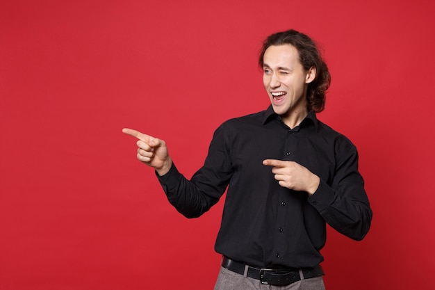 Knappe stijlvolle jonge krullend langharige man in zwart shirt poseren geïsoleerd op rode muur achtergrond studio portret. mensen oprechte emoties levensstijl concept mock up wijzende vinger hand op kopie ruimte