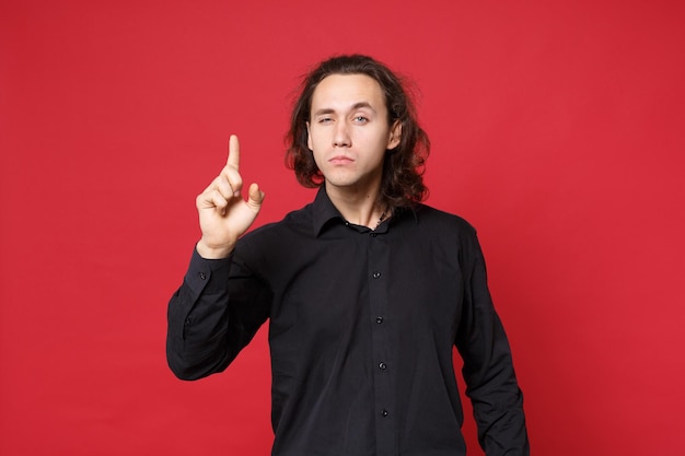 Knappe stijlvolle jonge krullend langharige man in zwart shirt poseren geïsoleerd op rode muur achtergrond studio portret. Mensen oprechte emoties levensstijl concept Mock up wijzende vinger hand op kopie ruimte